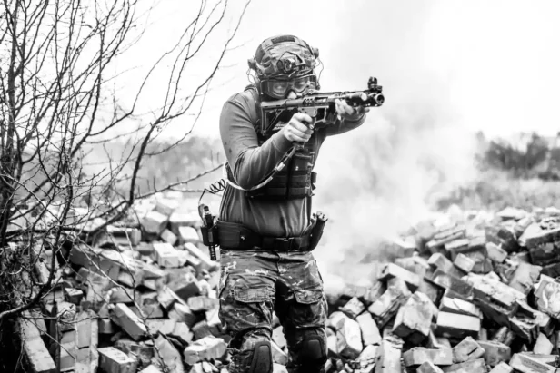 Man in tactical gear with rifle and body armor stands outdoors.